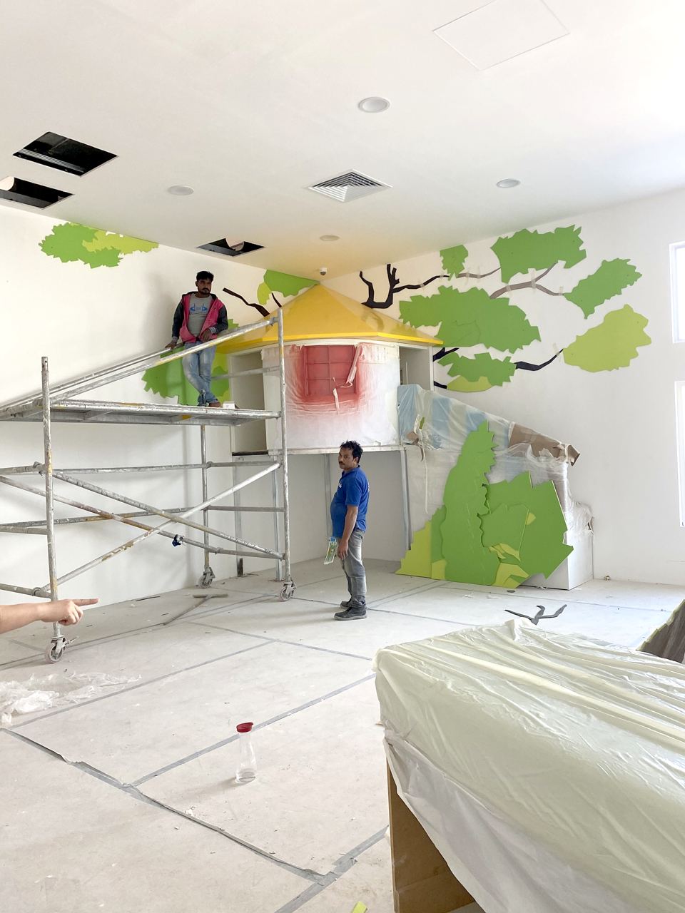 Installers fitting a treehouse and a tree canopy mural into a children's gallery.