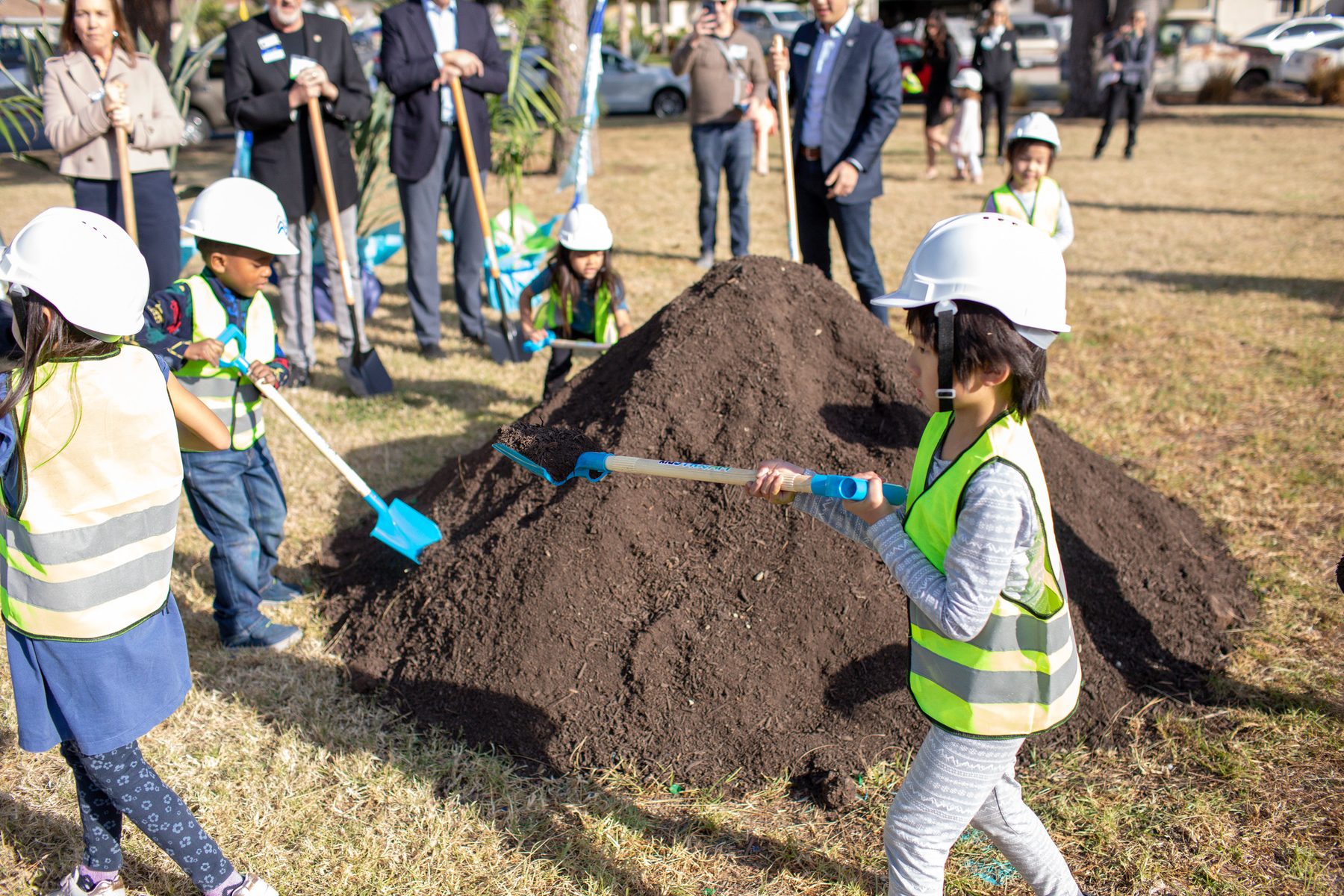 Children in hard hats and safety vests shoveling soil at a museum groundbreaking.
