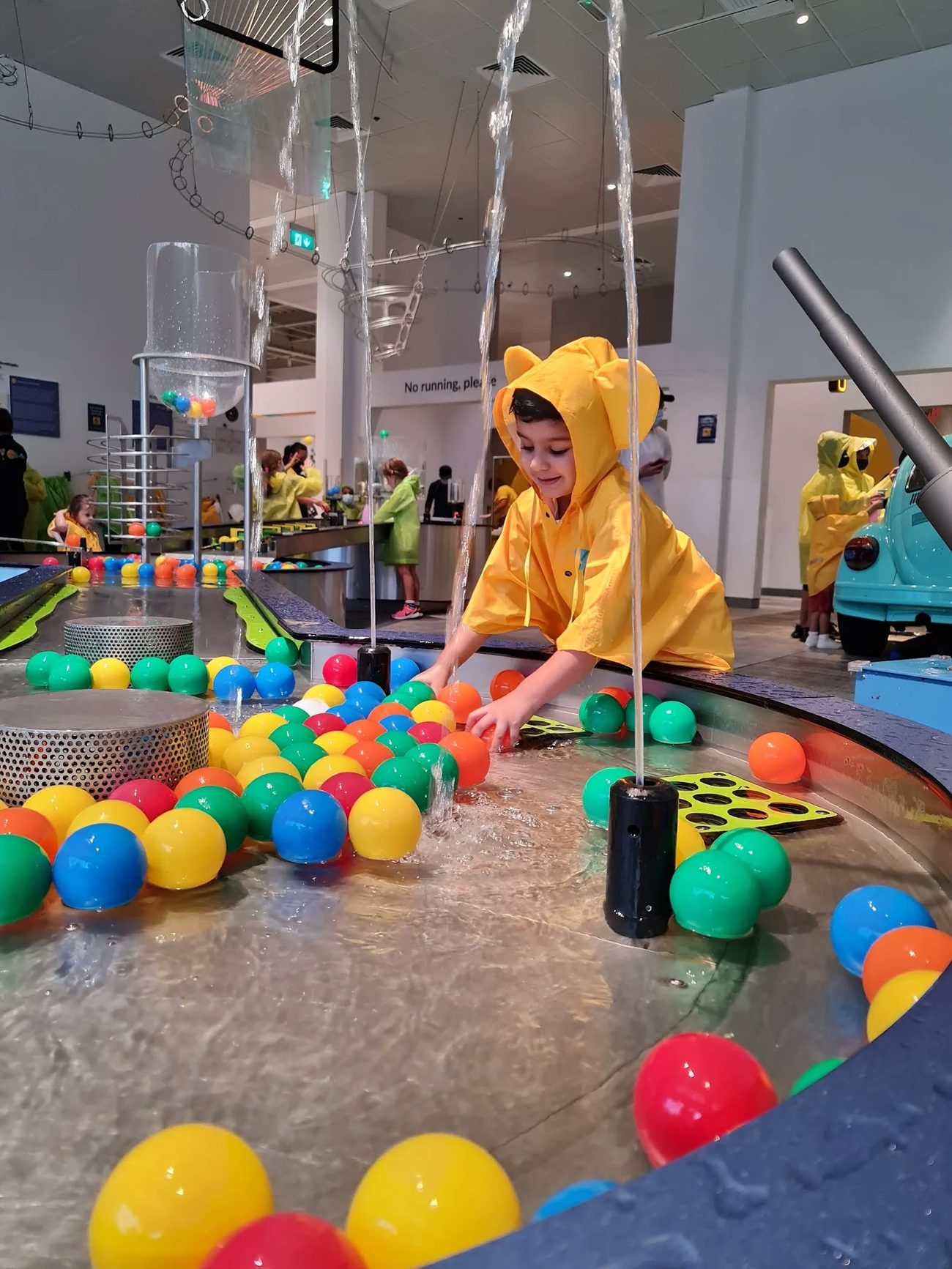 A child in a yellow raincoat playing at OliOli's colourful Water Gallery, surrounded by floating balls.