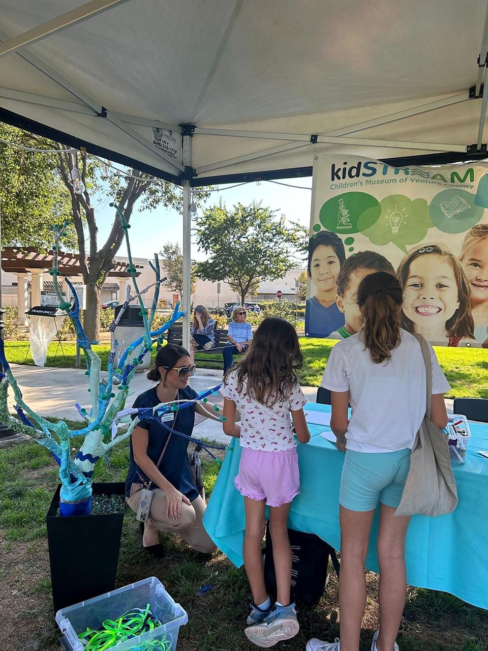 A kidSTREAM team member kneeling to help two girls at a community event booth.