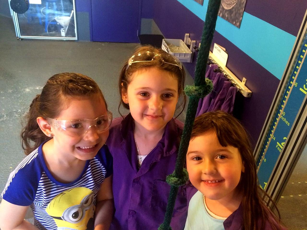 Three girls in goggles and purple lab coats smiling in a science gallery.