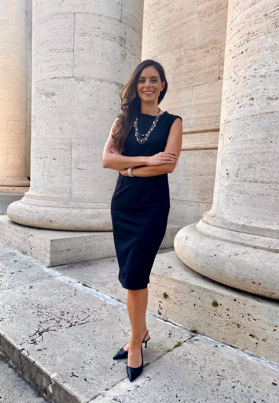 Dani Hildreth standing among tall classical stone columns, arms crossed.